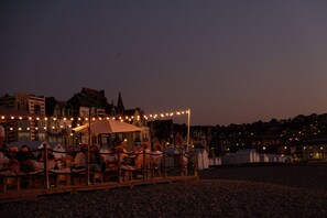 An outdoor seating area with tables and chairs, string lights overhead, and buildings in the background.