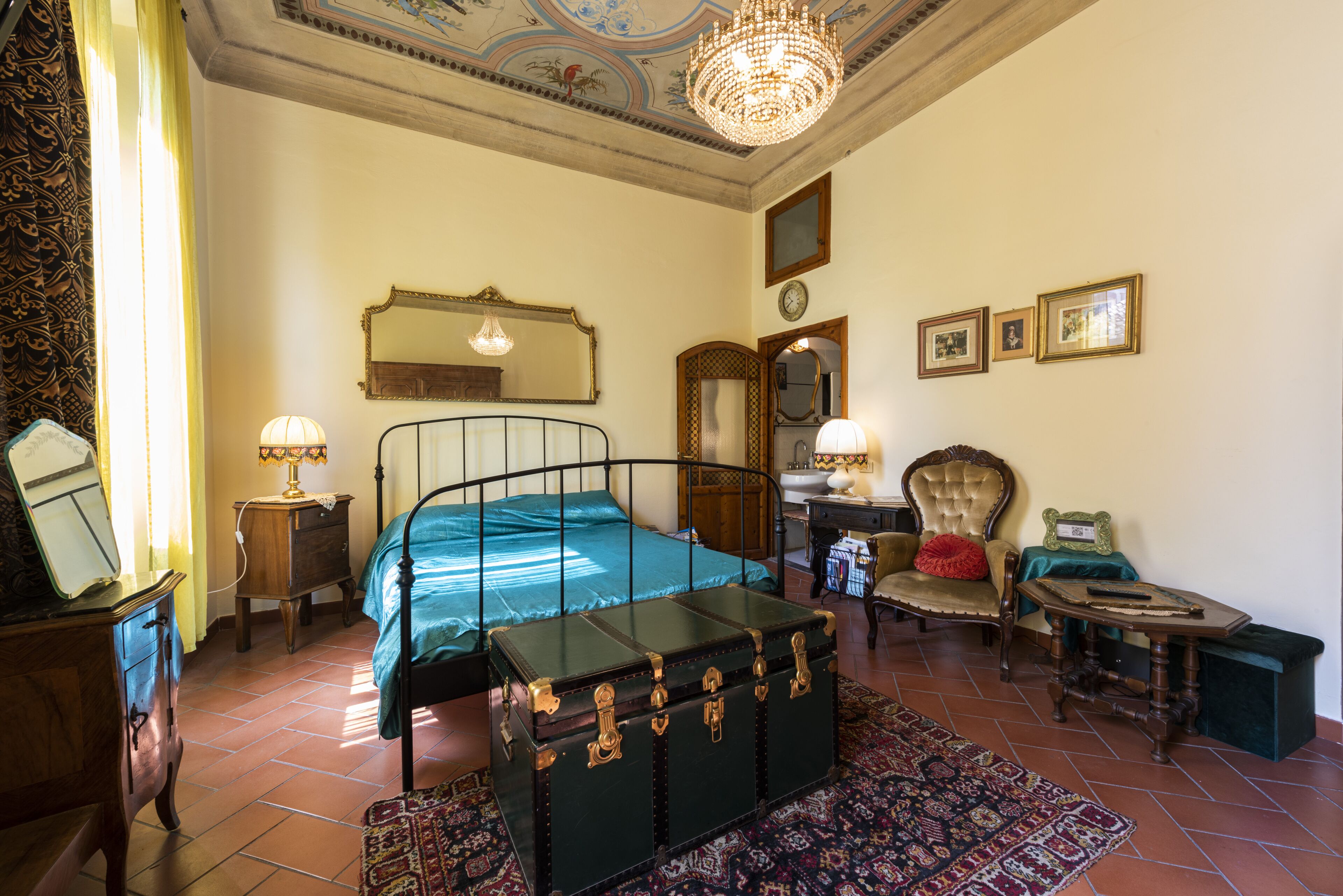A room with a bed, a chest of drawers, a chair, a table, a mirror, and a chandelier.
