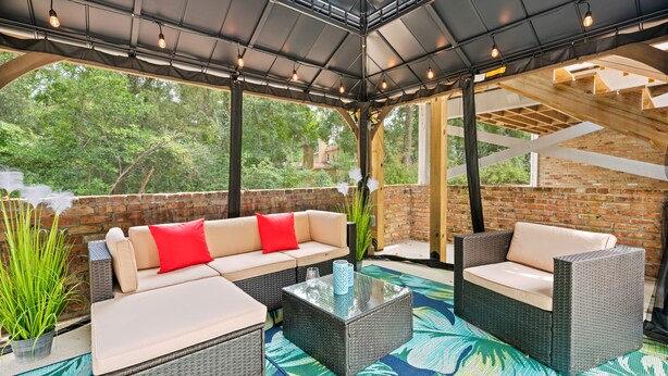 A covered patio area with wicker furniture, a glass coffee table, and a view of the surrounding greenery. - Airy 5br home w/pool, outdoor living, game room (Mobile)