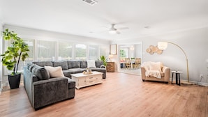 A spacious living room with a grey sectional sofa, a white coffee table, and a beige armchair. There is a dining area in the background with a table and chairs. - Airy 5br home w/pool, outdoor living, game room (Mobile)