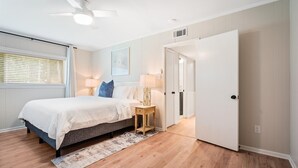 A bedroom with a bed, two bedside tables, a ceiling fan, a window with blinds, and a door. - Airy 5br home w/pool, outdoor living, game room (Mobile)