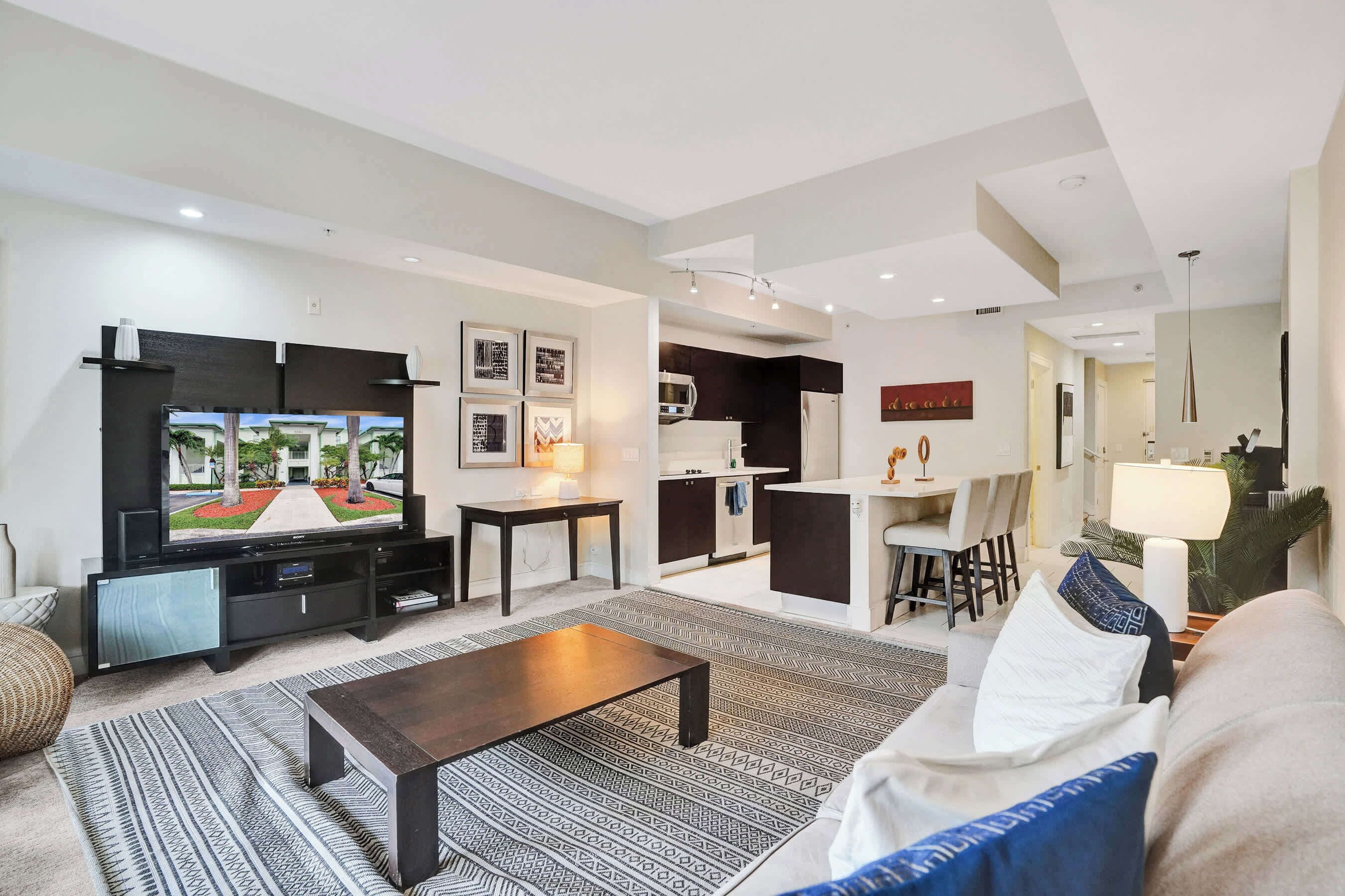 A modern living room with a flat-screen TV, a coffee table, and a sofa. There is a kitchen area in the background with a dining table and chairs.
