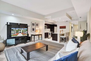 Uma sala de estar moderna com uma televisão de tela plana, uma mesa de centro e um sofá. É possível ver, ao fundo, uma área de cozinha com uma mesa de jantar e cadeiras.