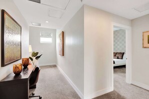 A hallway with a desk, chairs, and artwork on the walls. There is a door leading to a bedroom with a bed and pillows. - Villa Vista by VIAC - 2 Story w/ 3Bdrms & Golf View (Doral)