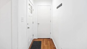 A narrow hallway with white walls and a wooden floor, leading to a white door with a small sign above it.