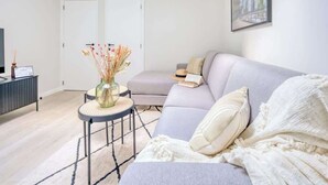 Una sala moderna con un sofá gris, una mesita redonda con un jarrón de flores secas, una estantería y un cuadro enmarcado en la pared.