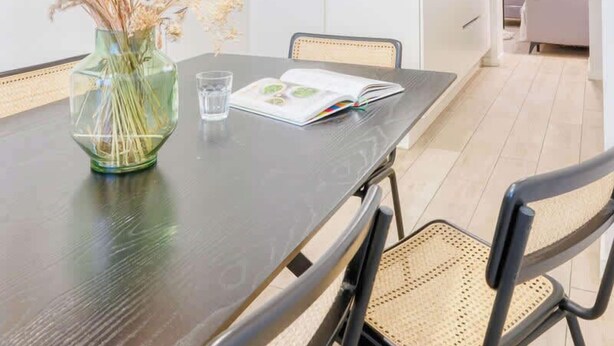 Una cocina moderna con armarios blancos, una mesa de comedor con sillas y un jarrón con flores secas.