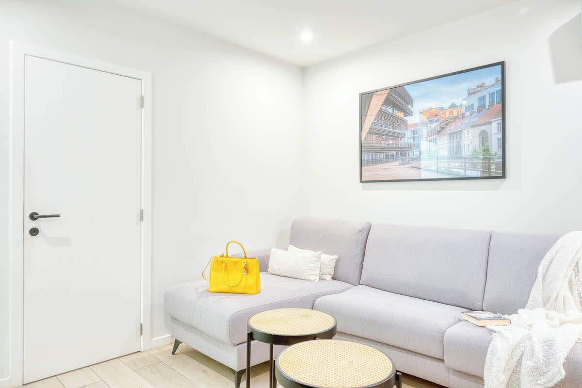 Un salon moderne avec un canapé gris, un sac à main jaune, deux tables rondes et un tableau encadré représentant un paysage urbain accroché au mur.
