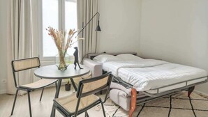 Ein kleiner, gut beleuchteter Raum mit einem Bett, einem runden Tisch mit Stühlen, einer Lampe und einer Vase mit getrockneten Blumen.