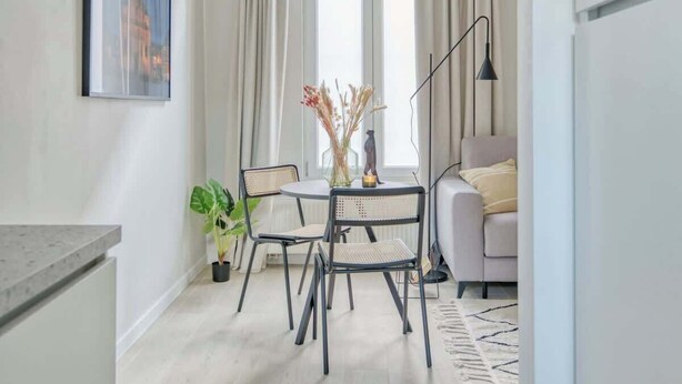 Eine moderne Küche mit Essbereich, ausgestattet mit einem runden Tisch und Stühlen, einem Blumentopf und Blick auf das Wohnzimmer mit Sofa und Vorhängen.