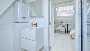 Moderni kylpyhuone, jossa on valkoinen pesuallas, peili ja kaappi. Suihkualueen yläpuolella on kattovalaisin, ja viereisessä huoneessa näkyy sänky.