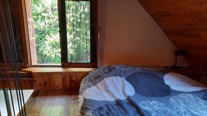 Ein Schlafzimmer mit Holzbalkendecke, einem großen Fenster mit Blick ins Grüne, einem Bett mit gemustertem Überwurf und einer hölzernen Fensterbank.