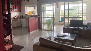 A spacious living area with a sofa, a coffee table, and a TV. There is a kitchen area with a refrigerator and a window. The room has a large sliding glass door leading to a balcony with a view of greenery.