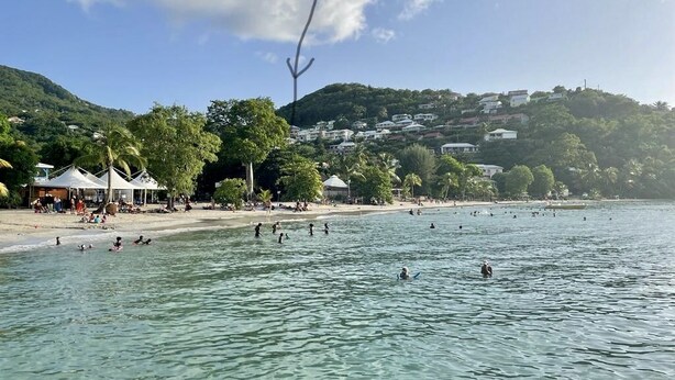 A beach with clear water, people swimming, and buildings on a hillside in the background.