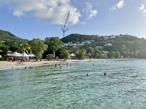En strand med klart vatten, folk som badar och byggnader på en sluttning i bakgrunden.