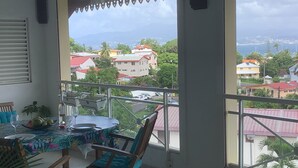 A balcony with a table and chairs, overlooking a residential area with houses and a view of the sea.