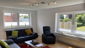 A modern living room with a dark blue sofa, a red rug, and a wooden coffee table. There are two windows with views of a brick wall and greenery outside. - Family vacation near the North Sea near Büsum. Great day trips with children on the agenda. (Heide)
