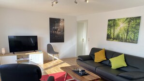 A modern living room with a flat-screen TV, a grey sofa with yellow cushions, a red rug, and a wooden coffee table. - Family vacation near the North Sea near Büsum. Great day trips with children on the agenda. (Heide)