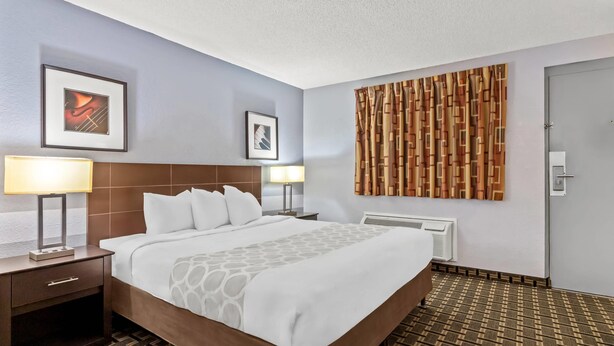 A hotel room with a large bed, two bedside tables with lamps, a wall-mounted artwork, a patterned curtain, and a radiator. - Rodeway Inn Albuquerque West (Albuquerque)