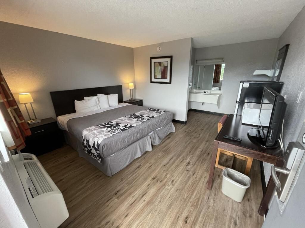 Habitación de hotel con cama, mesita de noche, lámpara, televisor y un pequeño escritorio con una silla.