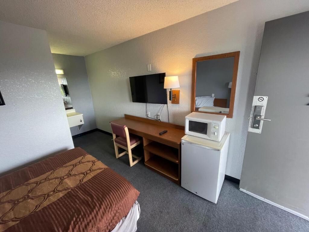 A hotel room with a bed, a desk, a chair, a television, a microwave, and a door with a lock.