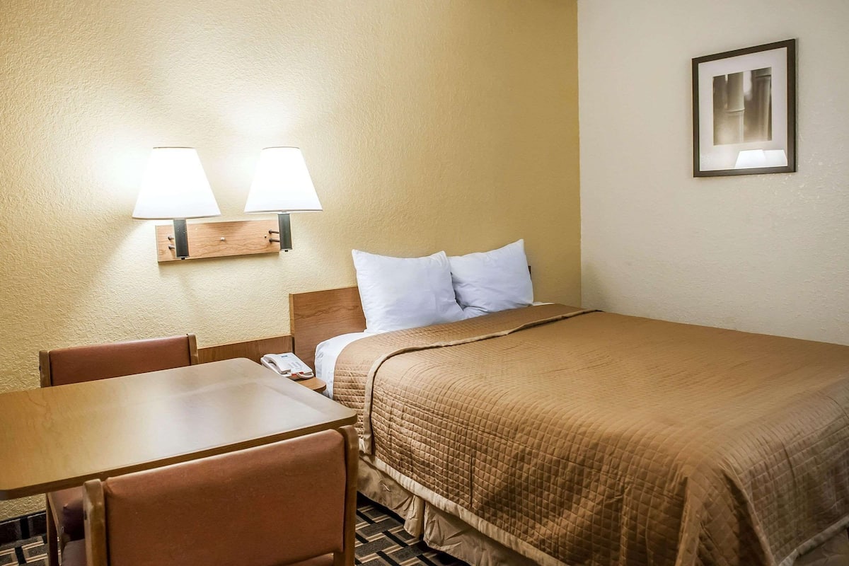 A hotel room with a bed, two bedside lamps, a wooden nightstand, a telephone, and a framed picture on the wall.