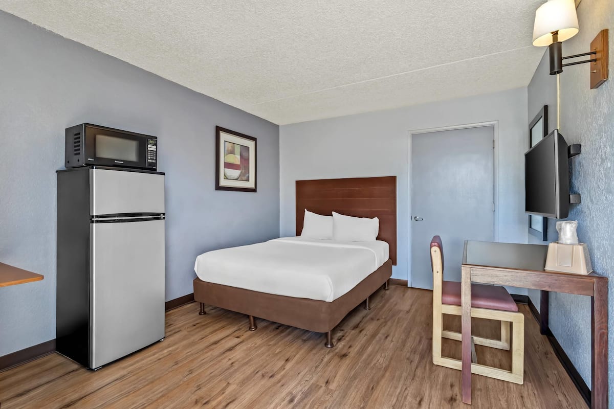 A compact hotel room with a bed, a small refrigerator, a microwave, a desk with a chair, and a television.