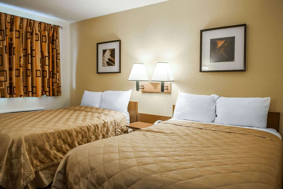 A hotel room with two beds, a window with patterned curtains, two wall-mounted lamps, and framed artwork on the walls.