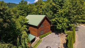 A log cabin with a green roof, surrounded by dense forest and a paved driveway. - Luxury cabin with private outdoor hot tub and access to resort pool (Sevierville)