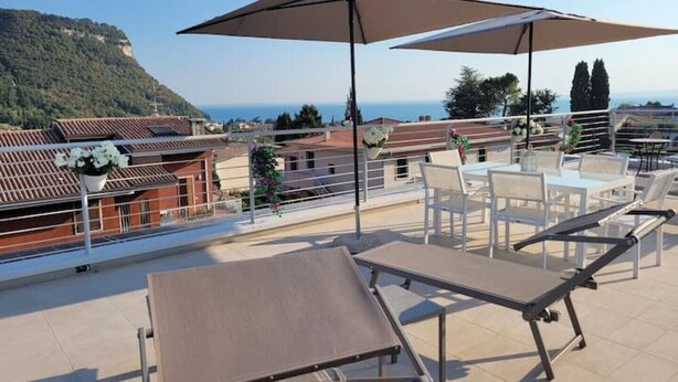 Une terrasse dotée de chaises longues et d’un grand parasol, offrant une vue sur une ville côtière aux toits de tuiles rouges et sur la mer.