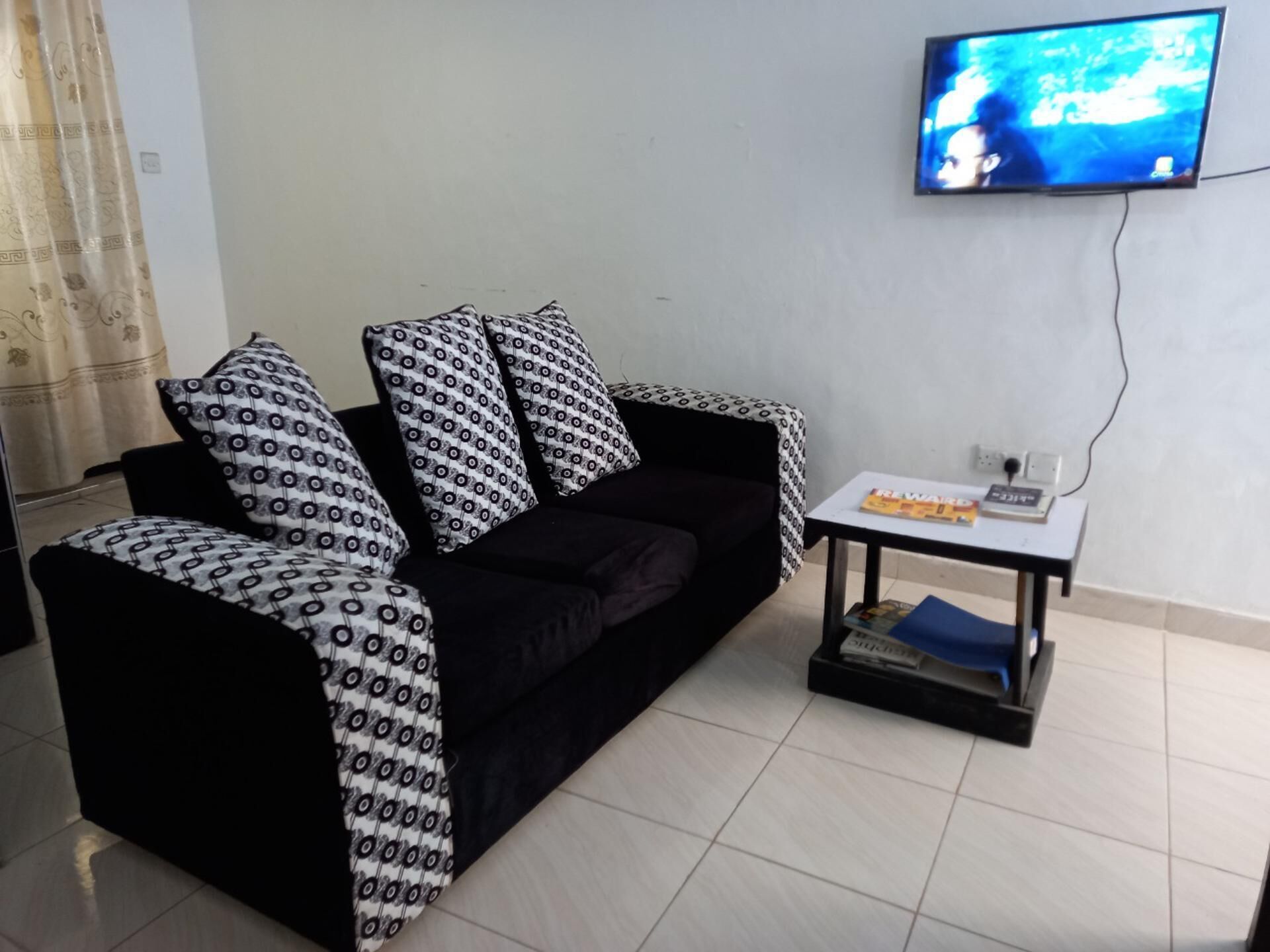 A living room with a black sofa, patterned throw pillows, a small table with a magazine, and a TV mounted on the wall.
