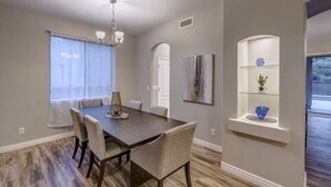 Un comedor con una mesa de madera oscura, sillas acolchadas, una araña y una ventana con cortinas traslúcidas.