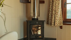 A room with a wood-burning stove, a wicker basket, a fire extinguisher, and a window with curtains. - Glamping in Wiltshire in our Luxury Shepherds Hut (Chippenham)