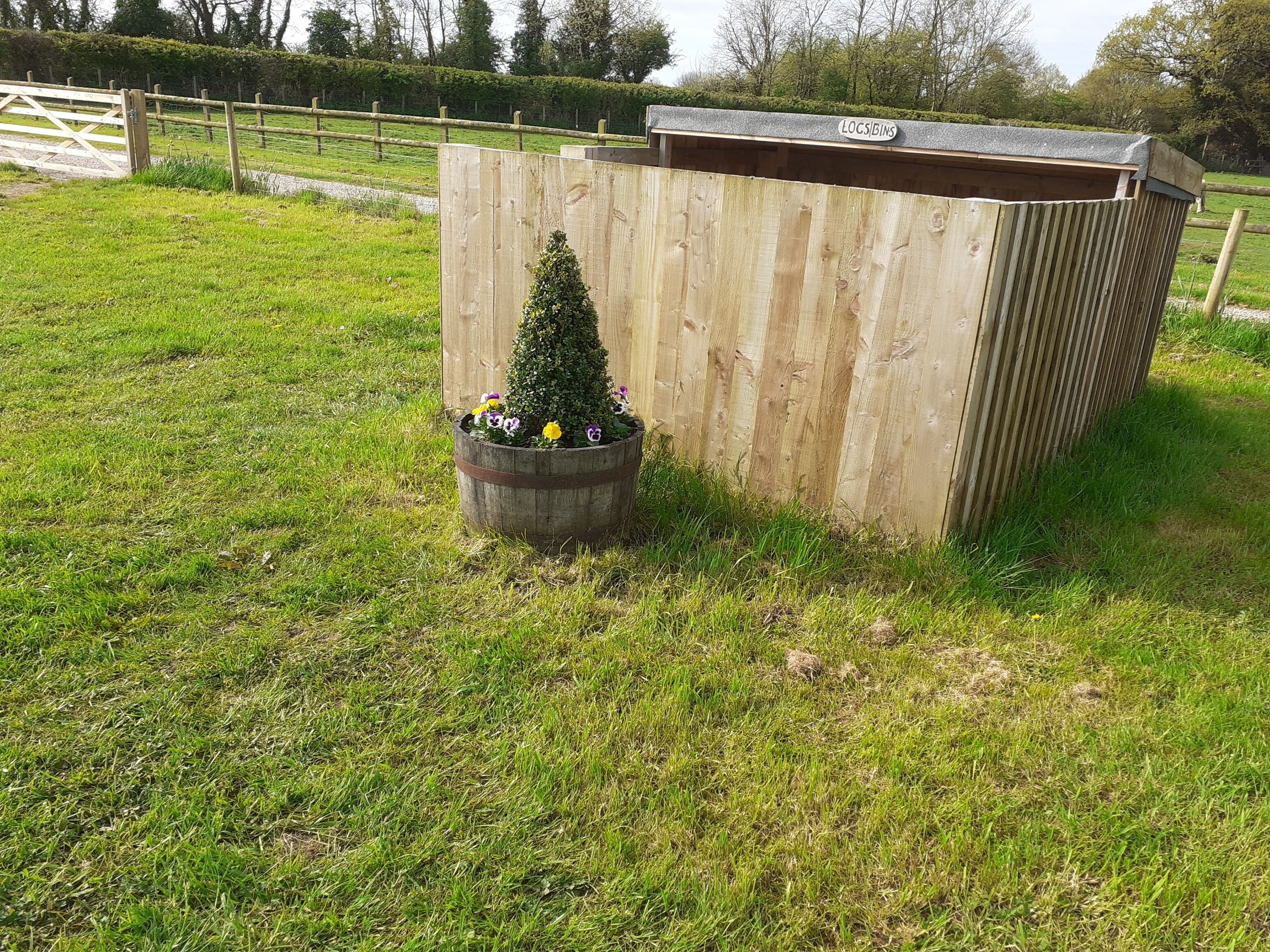 A wooden shed with a sign reading "LOCS BINS" in a grassy area with a wooden fence in the background.