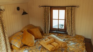 A bedroom with a bed, pillows, a window, curtains, and a lamp. - Glamping in Wiltshire in our Luxury Shepherds Hut (Chippenham)