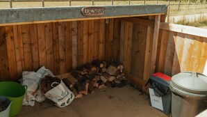 A wooden structure labeled "LOGS BINS" with a pile of firewood, a green bucket, and a trash bin labeled "DOG BAGS". - Glamping in Wiltshire in our Luxury Shepherds Hut (Chippenham)