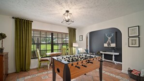 A room with a foosball table, a window with curtains, a chair, and a wall with framed pictures. - ‘Our Encanto’ -quiet golf course view-Tampa suburb (Valrico)