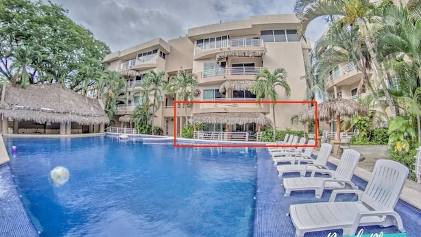 A swimming pool with lounge chairs and a thatched-roof structure, surrounded by palm trees and a multi-story building.