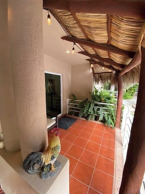 Una stanza con pavimento in piastrelle, tetto di paglia, una statua di un gallo e una porta che dà su un’area giardino con piante.