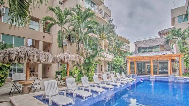 Una piscina con sillas de descanso y sombrillas, rodeada de palmeras y un edificio con balcones.