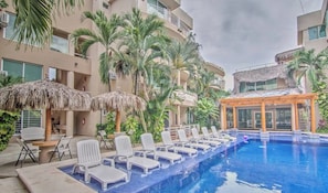 Una piscina con sedie a sdraio e ombrelloni, circondata da palme e da un edificio con balconi.