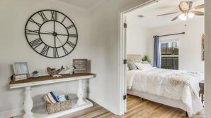 A bedroom with a bed, a large wall clock, a console table, and a sign that reads "WELCOME TO THE EAST HILL ESCAPE." - The East Hill Escape, A Bay View Beachy Cottage (Pensacola)