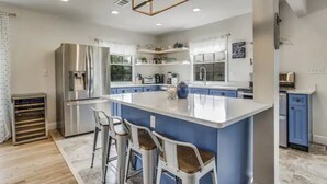 A modern kitchen with a large island, bar stools, a stainless steel refrigerator, and blue cabinetry. - The East Hill Escape, A Bay View Beachy Cottage (Pensacola)