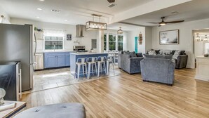 A modern kitchen with a central island, bar stools, and a grey sofa in a spacious living area with wooden flooring. - The East Hill Escape, A Bay View Beachy Cottage (Pensacola)