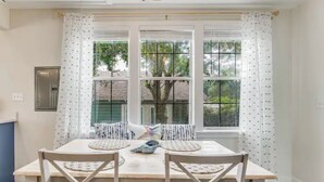 A dining area with a table and chairs, a window with curtains, and a view of trees outside. - The East Hill Escape, A Bay View Beachy Cottage (Pensacola)