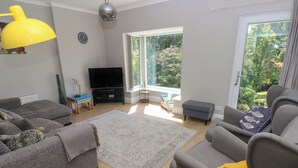 Ein Wohnzimmer mit einer Couch, einem Sessel und einem Fernseher. Ein Fenster bietet Blick auf Grünflächen.