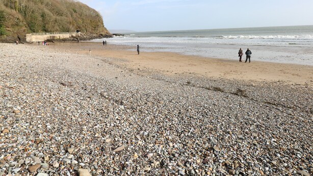 Ein Kiesstrand mit Menschen, die darauf spazieren gehen, und einer Brücke im Hintergrund.