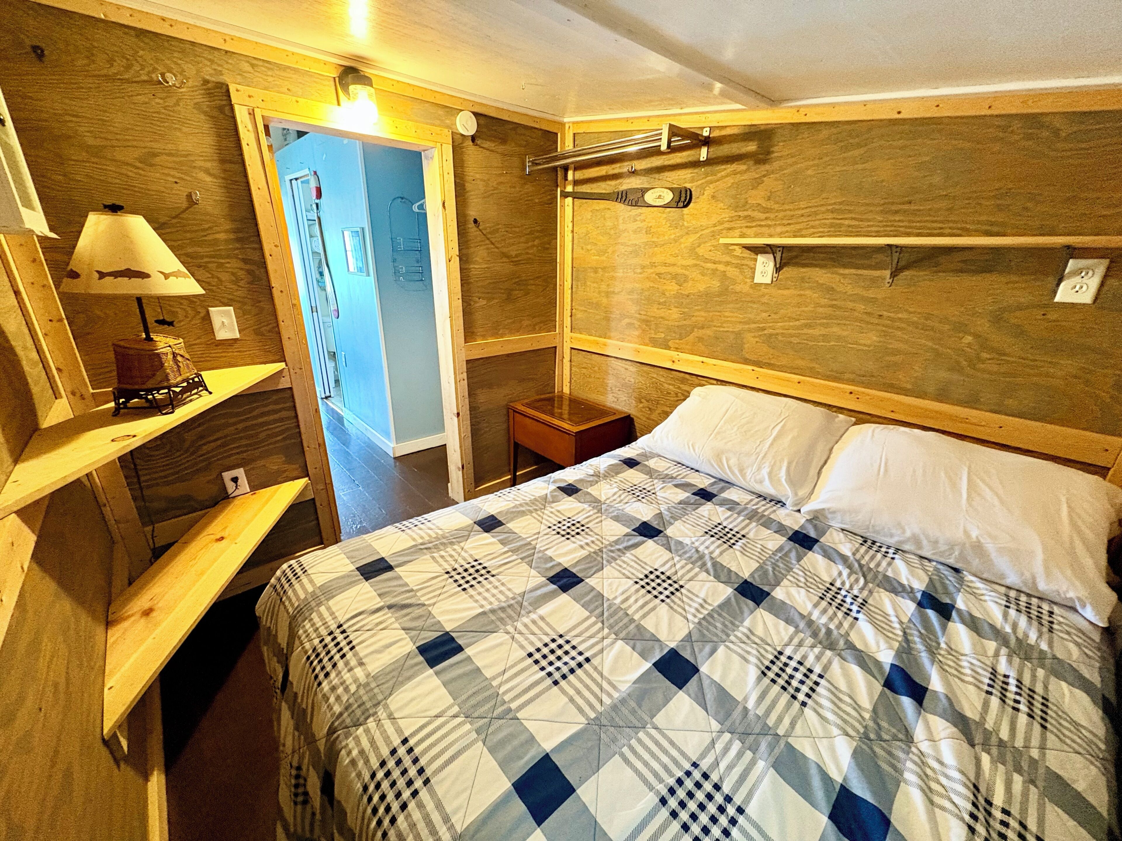 A wooden room with a bed, a nightstand, a lamp, and a shelf with a basket.