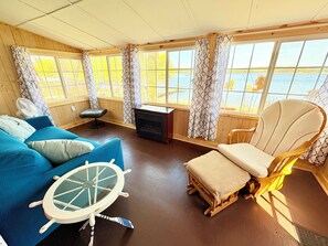 A room with a blue sofa, a wooden chair, a small table with a ship wheel, and large windows overlooking a body of water. - Lakefront Cabin w/ Dock & Kayaks, Perfect Summer Stay
Rustic Nautic - Cabin 9 (South Haven)