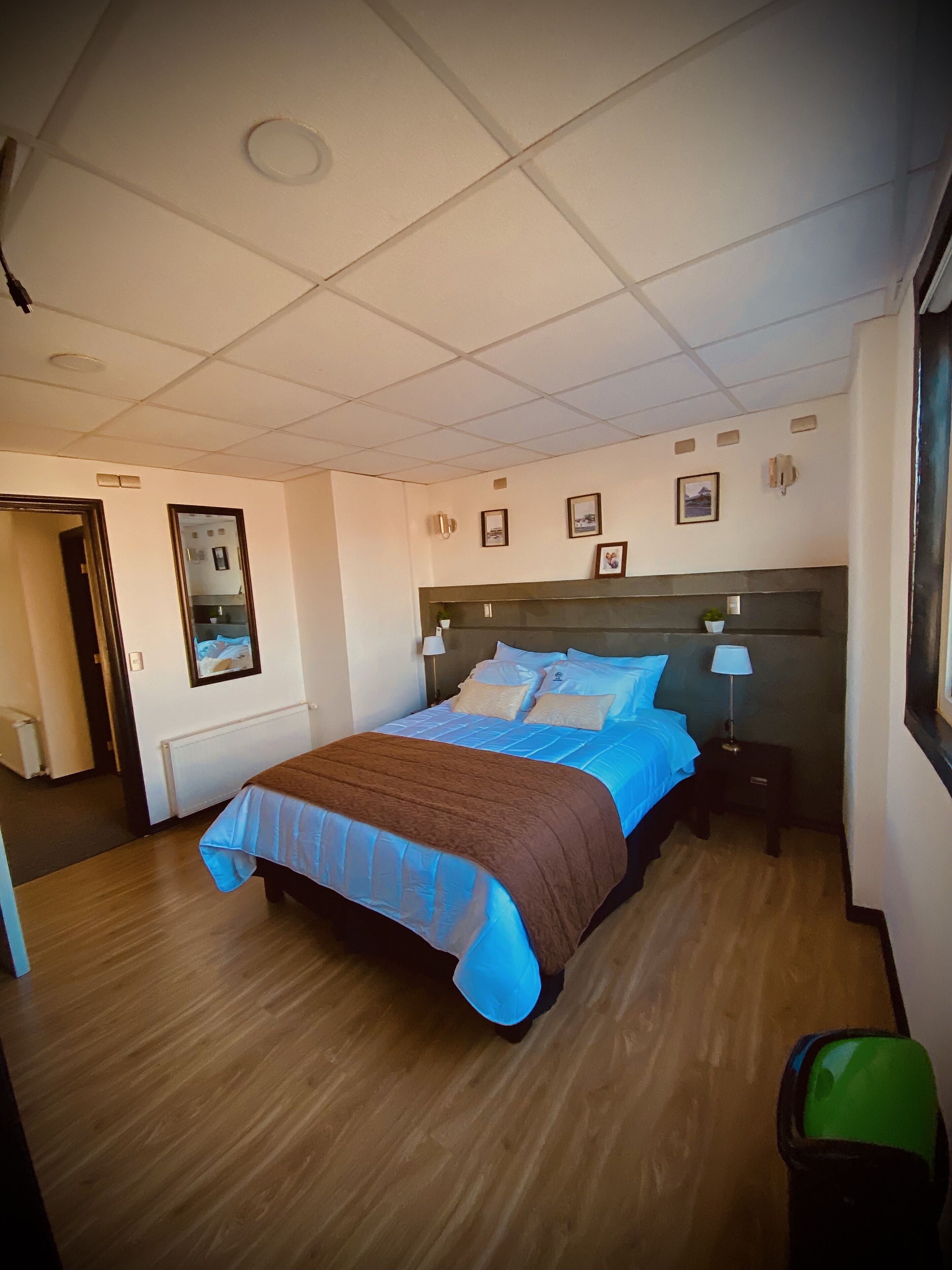 Une chambre d’hôtel avec un grand lit, une table de chevet avec une lampe, une petite table de chevet avec une lampe, une poubelle verte et une porte donnant sur une autre pièce.